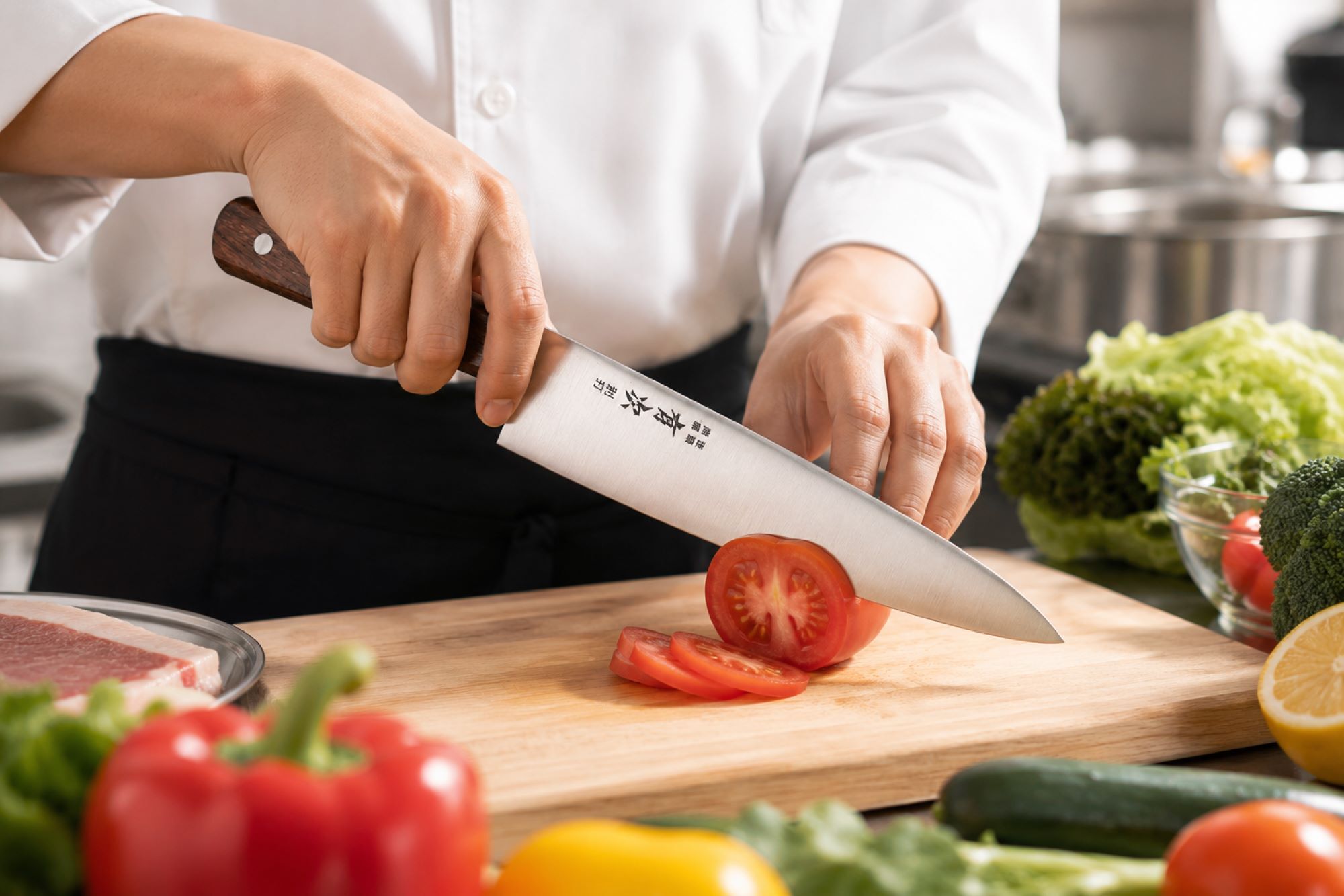 A professional chef's hands using a Japanese Gyuto chef's knife to slice a fresh heirloom tomato on a wooden cutting board in a bustling restaurant kitchen background.